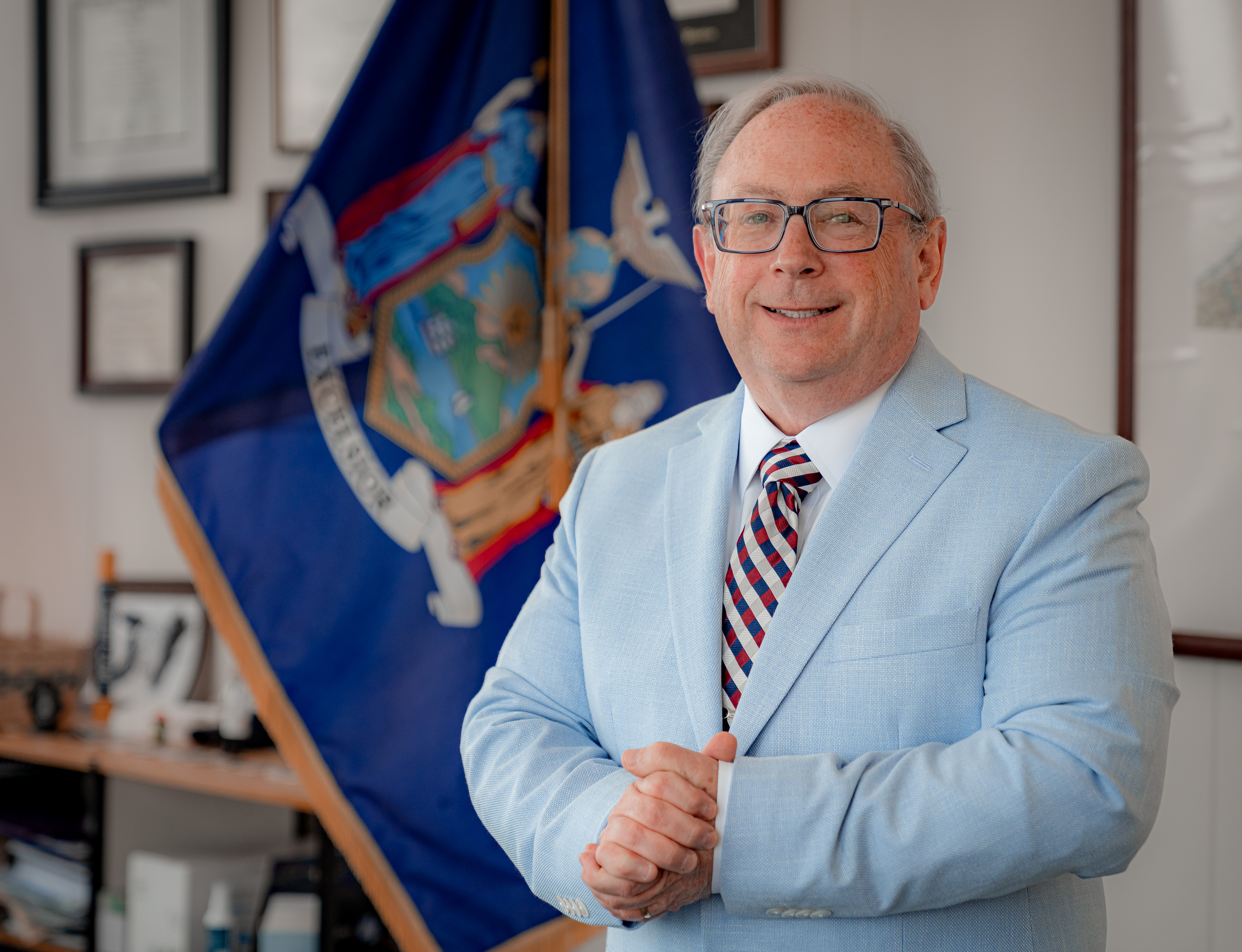 An older white man posing with a smile in front o a New York State flag in an office. He has gray hair and his hands are folded over his chest.