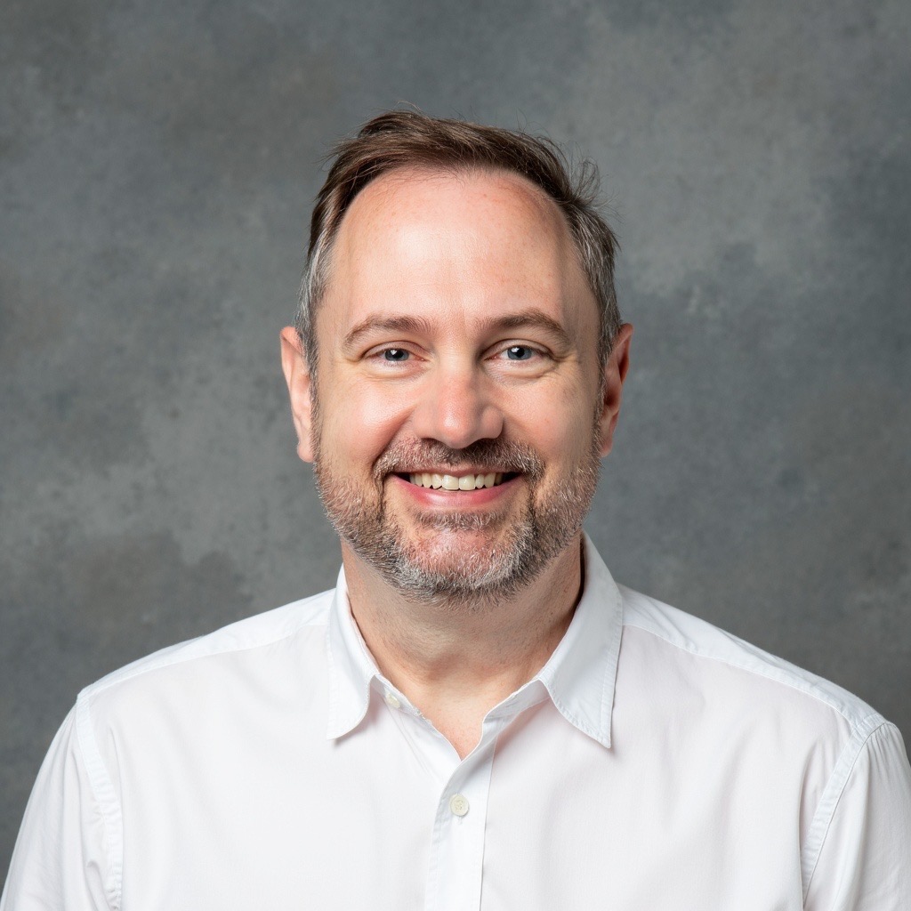 A middle aged white main with a salt and pepper beard smiles at the camera in the manner of a headshot