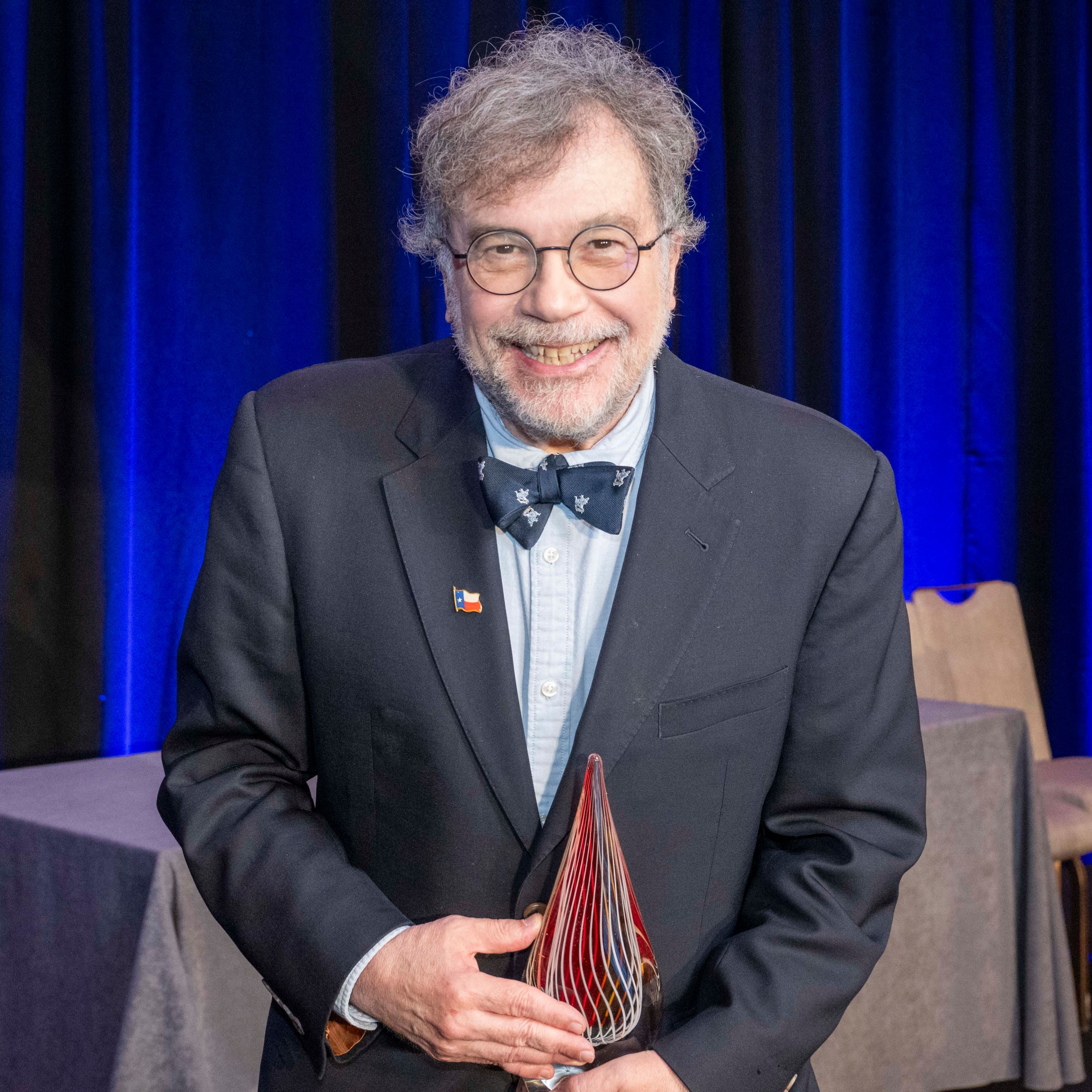 A man with round glasses, a shaggy gray beard and gray hair holds an award while smiling