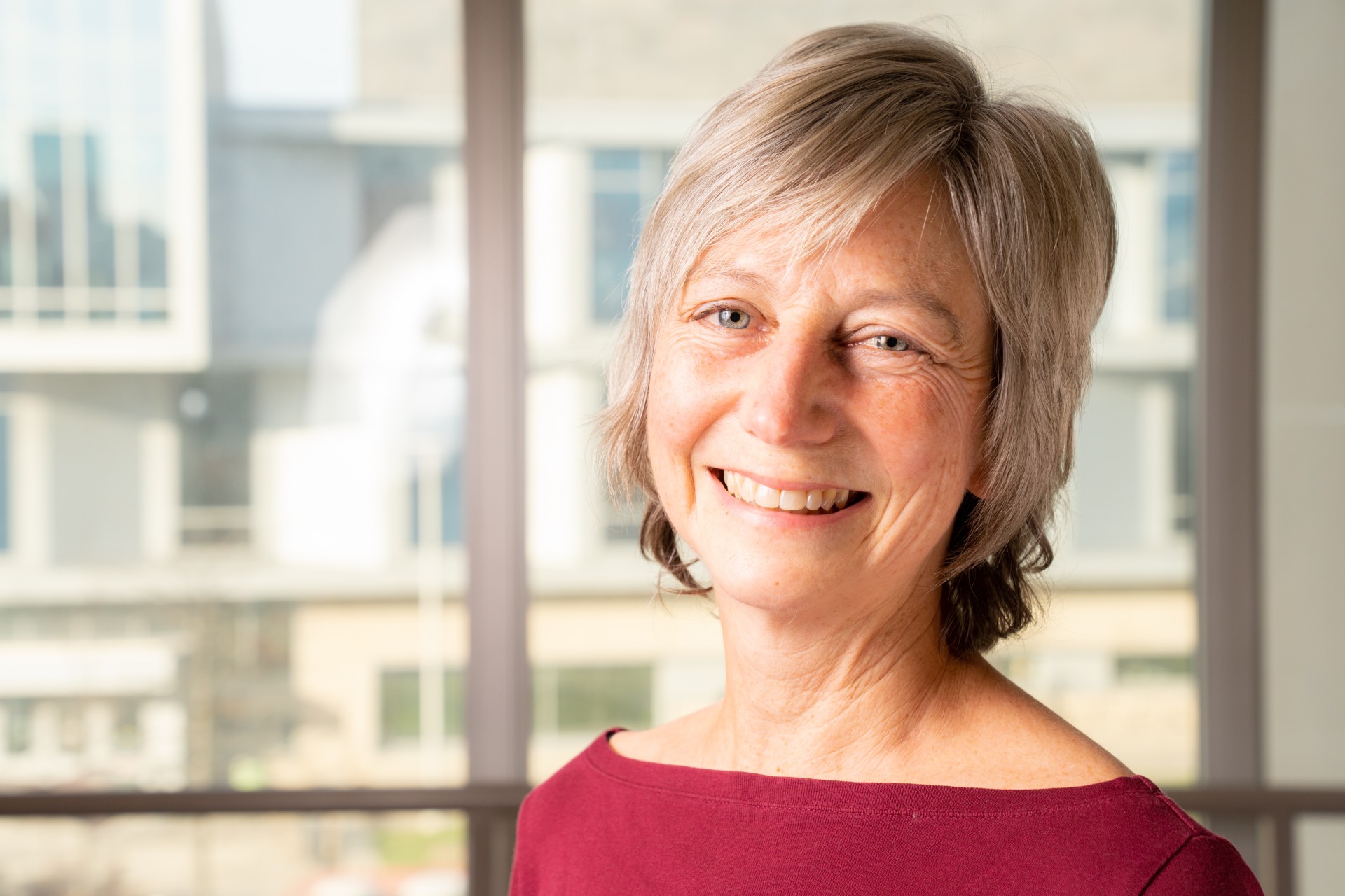 An older white woman with shorter blonde hair tilts her head and smiles at the camera